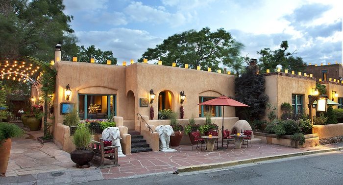 A rustic adobe-style restaurant features outdoor seating with red umbrellas amidst vibrant plants, situated on a cobblestone sidewalk under a partly cloudy sky.