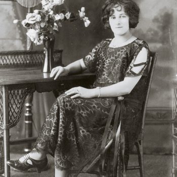 A woman sits elegantly in a patterned dress, posing beside a table with flowers and a lamp, against a vintage studio backdrop.