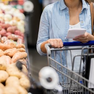 Person holds shopping cart and list, standing beside a produce section filled with various potatoes in a grocery store.