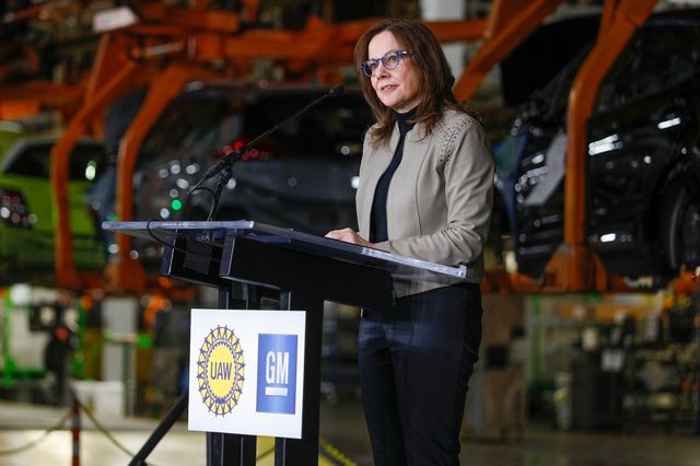 Woman speaks at podium with UAW and GM logos; cars on assembly line background.