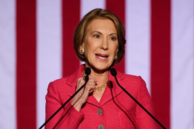 A woman speaks at a podium, wearing a pink jacket, against a backdrop of red and white vertical stripes.