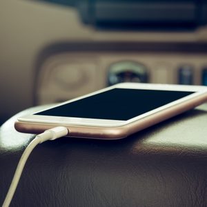 Smartphone charging on a car console, connected with a white cable. Dashboard controls visible in the background, creating an automotive setting.