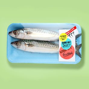 Two fish lie in a blue tray with expiration stickers showing various dates, set against a pale green background.