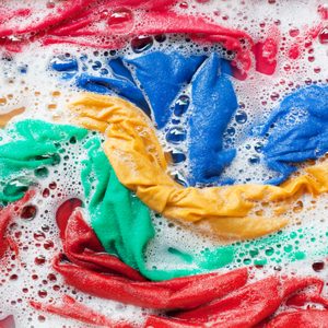 Colorful clothes soak in soapy water with bubbles covering the vibrant fabric; red, blue, yellow, and green cloths are visible.
