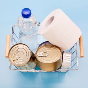 Basket holds bottled water, canned food, and a toilet paper roll on a blue background.