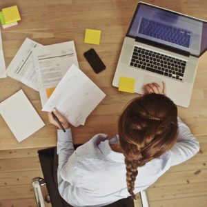 Person working on a laptop at a wooden desk, surrounded by papers, sticky notes, a smartphone, and a glass of water.