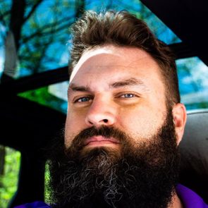 Bearded man gazes forward, sunlight casting shadows, seated inside a vehicle with a view of trees through a sunroof.