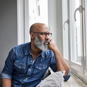 A man with a beard looks out a window, resting his chin on his hand in a contemplative pose, wearing a denim shirt indoors.