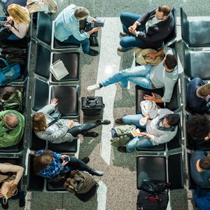 People sit on black chairs using phones and reading in an airport waiting area, surrounded by luggage and bags on the tiled floor.