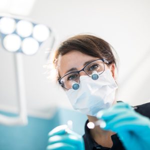 Dentist wearing magnifying glasses and mask, holding dental tools, in a brightly lit examination room.