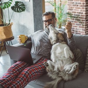 Man relaxing with dog on couch, holding a mug, using a laptop. Comfortable home setting with plants and wooden accents.