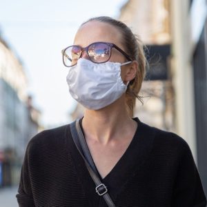 Person wears sunglasses and a white mask, walking on a sunny street with blurred buildings in the background.