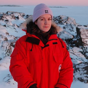 Person in red parka stands in snowy, rocky landscape during sunset, wearing a purple hat with an embroidered logo.
