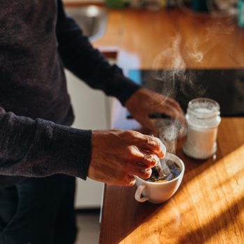 Hands stir steaming coffee in a white mug on a wooden kitchen counter, next to a jar of sugar.