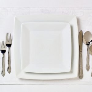 Square plates are centered on a white placemat, surrounded by forks on the left and a knife, spoon, and teaspoon on the right.