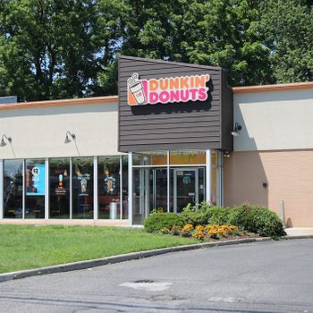 A Dunkin' Donuts shop with glass windows displays promotional posters; surrounded by greenery and a parking area under clear skies.