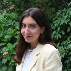 A person smiles outdoors, wearing a yellow jacket and necklace, surrounded by lush green foliage.