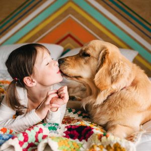 Child kisses golden retriever on a bed, under a colorful, crocheted blanket; geometric wall pattern in background.