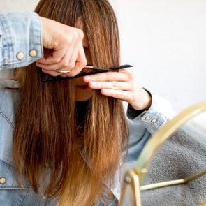 Person trims their bangs with scissors, using a comb for guidance, in front of a mirror, wearing a denim shirt.