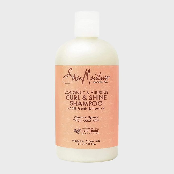 Bottle stands containing SheaMoisture Curl & Shine Shampoo, featuring coconut, hibiscus, silk protein, neem oil; designed for thick, curly hair. Cream cap, peach label.