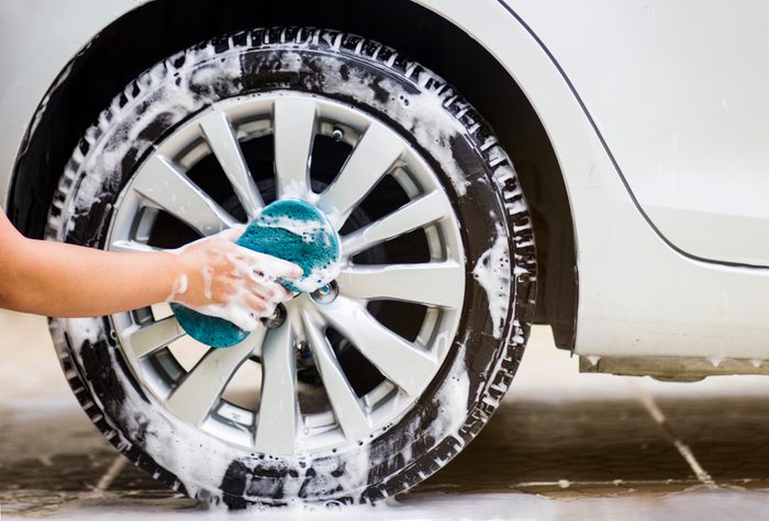 A hand scrubs a car wheel with a soapy sponge, in a vehicle cleaning setting.