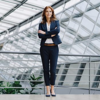 A woman stands confidently with crossed arms, wearing a suit, in a modern glass-roofed building with metal railings and plants.