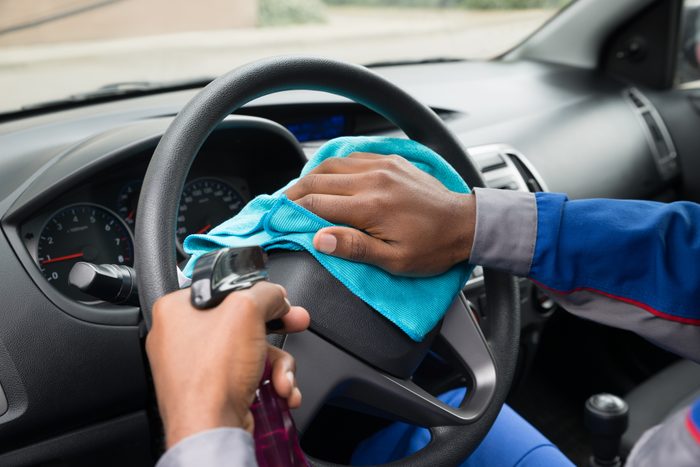 Hands clean a steering wheel using a blue cloth and spray bottle inside a parked car.