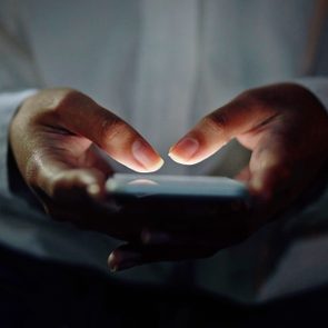 Hands typing on a smartphone, glowing in the dark, creating an illuminated contrast against a dim background.