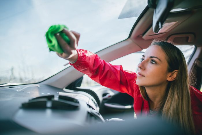 Person wipes car windshield with green cloth, focused expression, wearing red jacket, inside a vehicle with a clear sky visible through the glass.