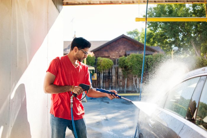 Man pressure-washes car, wearing a red shirt, in a sunny outdoor carwash. Background features trees and a wooden fence.