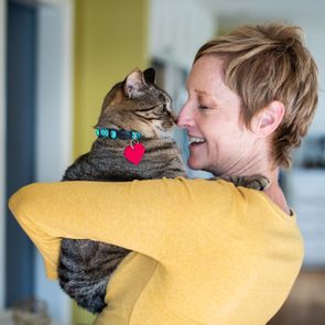 Woman holding a cat nuzzling her face, inside a warmly lit home. The cat wears a collar with a red heart tag.