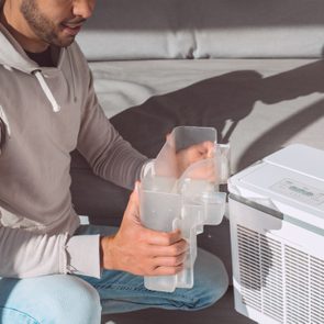 Person holding a transparent water tank, kneeling beside a dehumidifier, in a living room with a gray couch and natural lighting.