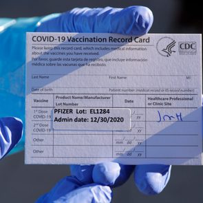 Gloved hand holds COVID-19 vaccination card; displays vaccine type as Pfizer with date: 12/30/2020. Background shows a blurred blue protective garment.