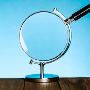 Magnifying glass held by stand, tilted upward, against a wooden surface and bright blue background.