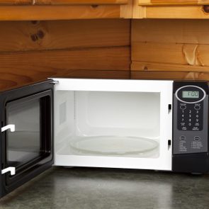 Microwave sits open on a green countertop, displaying a timer set to 1:00, in a wooden kitchen corner with visible electrical outlet.