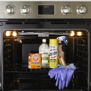An oven holds cleaning supplies, including baking soda, vinegar, cleaner spray, gloves, and a brush, under bright interior lights in a kitchen setting.