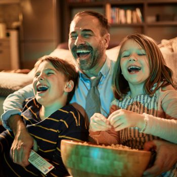 A man and two children are laughing on a sofa, holding popcorn and a remote, in a cozy living room.