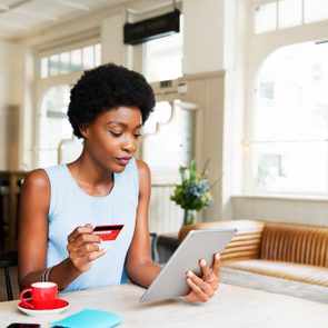 A person holds a credit card and tablet, sitting in a bright, cozy cafÃ© with a wooden table and red cup.