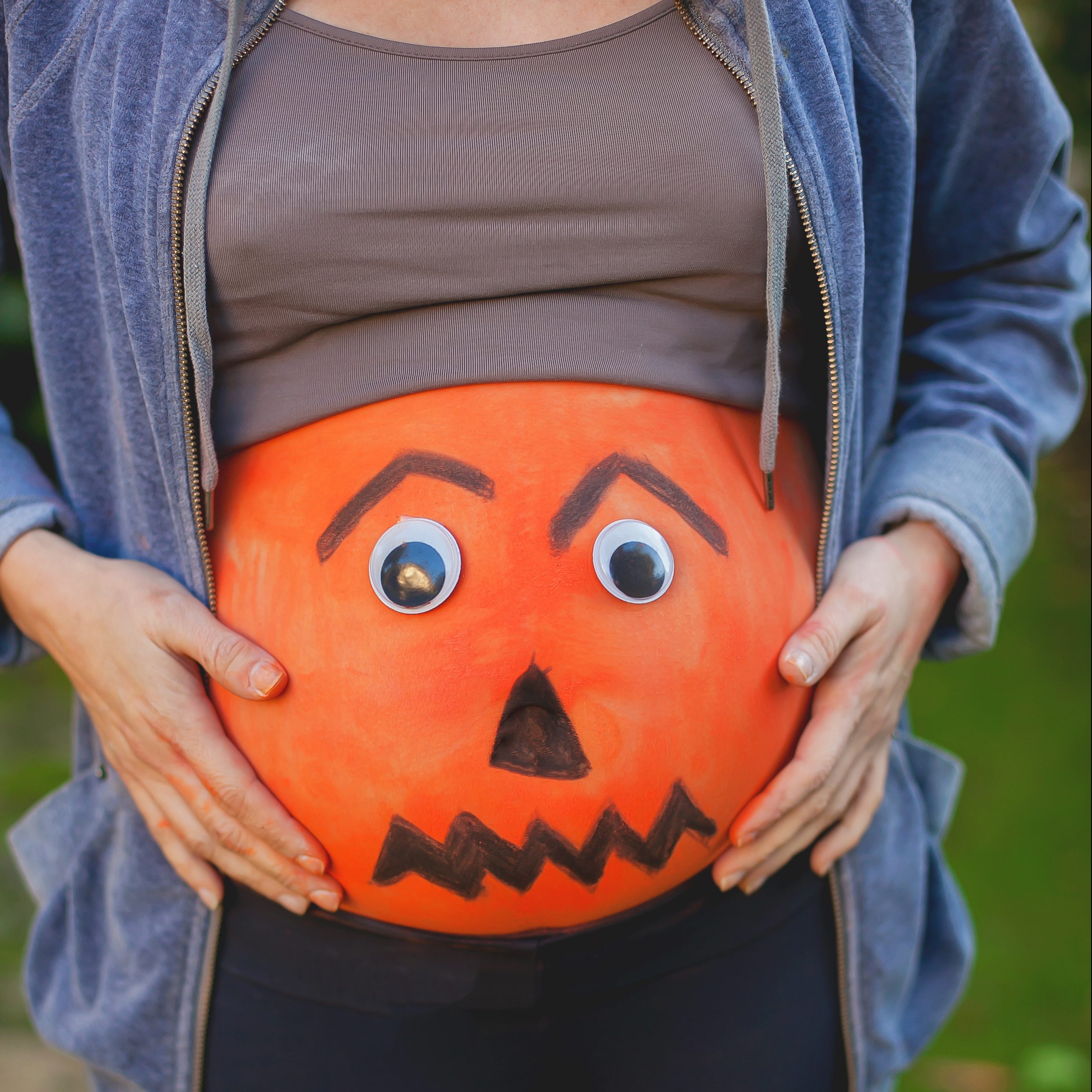 Pregnant belly painted like a jack-o'-lantern with googly eyes, held by hands, outdoors.