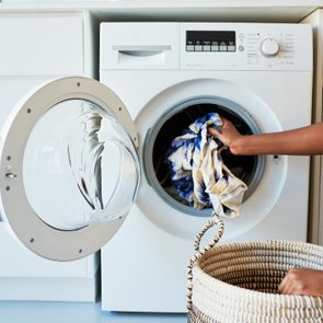 A person places patterned clothes into a front-loading washing machine, with a woven basket nearby in a laundry room.