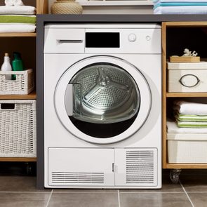 A front-loading washing machine sits unused in a laundry room with shelves holding baskets, folded towels, and cleaning supplies.