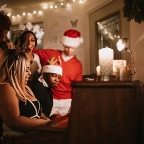 People sing around a piano decorated with candles and holiday lights, wearing festive attire in a cozy room.