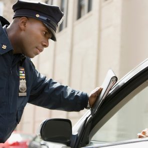 Police officer leans on car window, engaging with female driver, urban street backdrop.