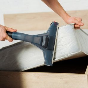 Vacuum cleaner suctioning dirty mattress, lifted by a person, in a bedroom setting with a wooden bed frame.