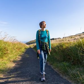 A person walks joyfully along a paved path, surrounded by tall grasses under a clear blue sky.