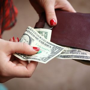 Hands hold a leather wallet, pulling out U.S. dollar bills; background features blurred plaid clothing.