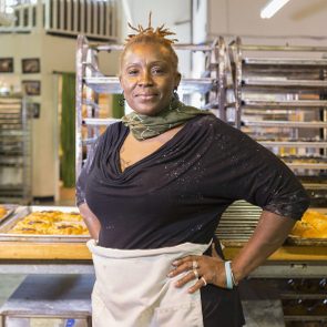 A baker stands confidently with hands on hips, surrounded by trays of freshly baked bread in an industrial kitchen.