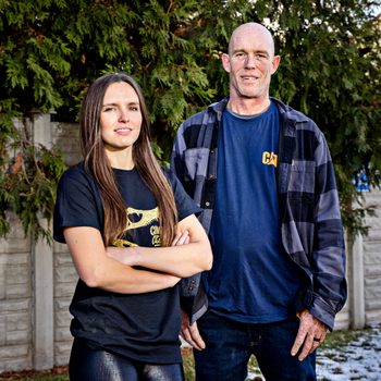 Two people stand confidently outside, surrounded by greenery and a concrete fence, wearing casual shirts.