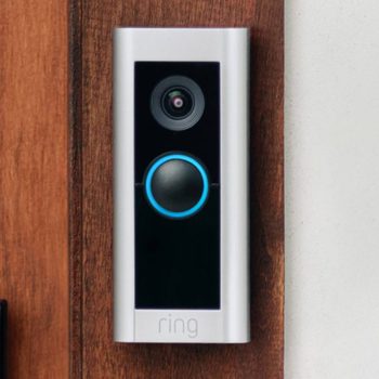Doorbell camera displays a blue-lit button and lens on a wooden wall. "Ring" text is at the bottom.