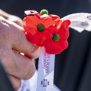 Hand holding red poppy flowers with green centers, attached to white ribbon reading 